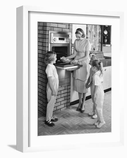 1950s MOTHER AND THREE DAUGHTERS STANDING AROUND OVEN IN KITCHEN BAKING PIE-Panoramic Images-Framed Photographic Print