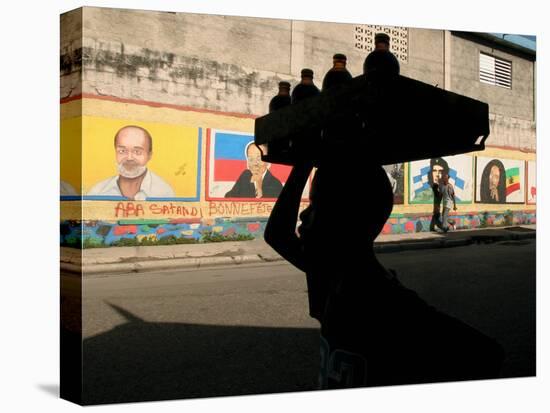 A Boy Carrying Bottles on His Head Passes by a Wall with Pictures of Haitian President Renel Preval-Ariana Cubillos-Stretched Canvas