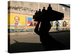 A Boy Carrying Bottles on His Head Passes by a Wall with Pictures of Haitian President Renel Preval-Ariana Cubillos-Stretched Canvas