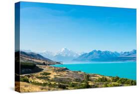A road winds along the edge of a turquoise blue lake with mountains in the distance, New Zealand-Logan Brown-Stretched Canvas