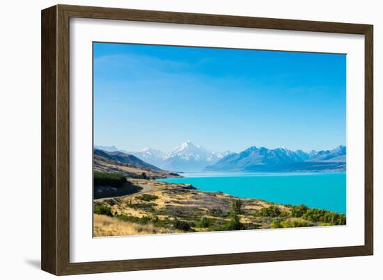 A road winds along the edge of a turquoise blue lake with mountains in the distance, New Zealand-Logan Brown-Framed Photographic Print