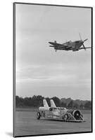 Airplane Flying over Race Car in Action-null-Mounted Photographic Print