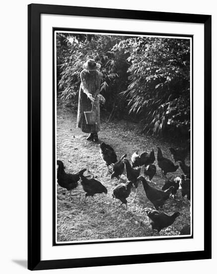 An Older Woman in a Long Dress and Wide-Brimmed Hat Throws Handfuls of Chicken Feed-null-Framed Art Print