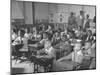 Children Sitting at their Desks in a Classroom, Teachers at the Rear of the Room-null-Mounted Photographic Print