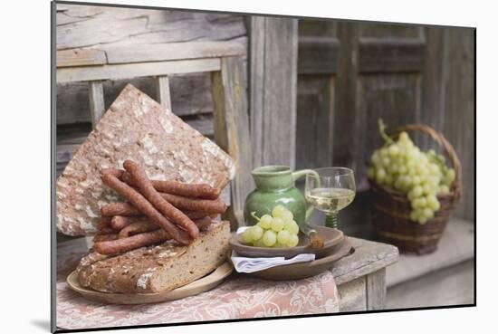 Grapes, Bread, Sausages and Wine on Wooden Bench in Front of Farmhouse-Eising Studio - Food Photo and Video-Mounted Photographic Print