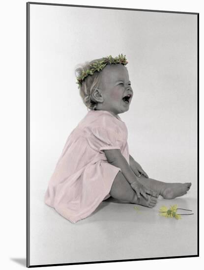 Little Girl Sitting and Laughing with a Floral Ring on Head and Two Flowers in Front of Her-Nora Hernandez-Mounted Giclee Print