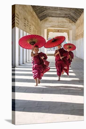 Myanmar, Mandalay Division, Bagan. Three Novice Monks Running with Red Umbrellas in a Walkway (Mr)-Matteo Colombo-Stretched Canvas