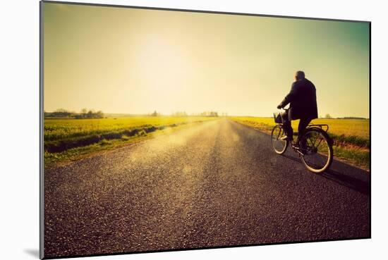 Old Man Riding A Bike On Asphalt Road Towards The Sunny Sunset Sky-Michal Bednarek-Mounted Art Print