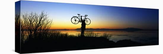 Silhouetted Cyclist Holding Bicycle over Head, River's Edge, Sunset, Alaska-null-Stretched Canvas