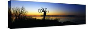 Silhouetted Cyclist Holding Bicycle over Head, River's Edge, Sunset, Alaska-null-Stretched Canvas