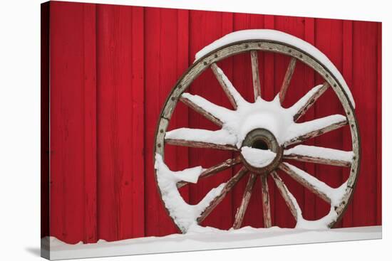 Snow-covered wagon wheels against red barn near town of Banff, Canadian Rockies, Alberta, Canada-Stuart Westmorland-Stretched Canvas