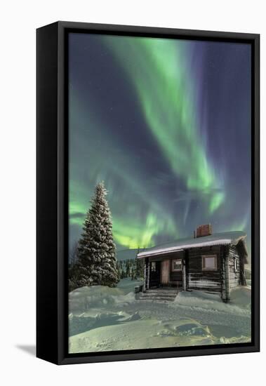 The Northern Lights (Aurora borealis) frame the wooden hut in the snowy woods, Pallas, Yllastunturi-Roberto Moiola-Framed Stretched Canvas