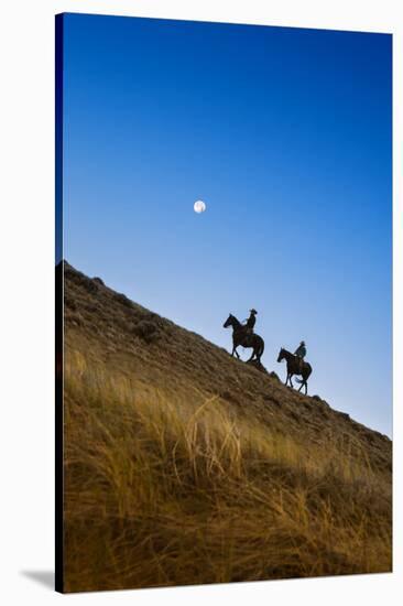 Two wranglers riding horses up a hill with full moon in background at blue hour-Sheila Haddad-Stretched Canvas