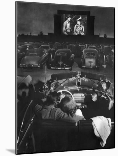 Young Couple Snuggling in Convertible as They Watch Large Screen Action at a Drive-In Movie Theater-J^ R^ Eyerman-Mounted Photographic Print